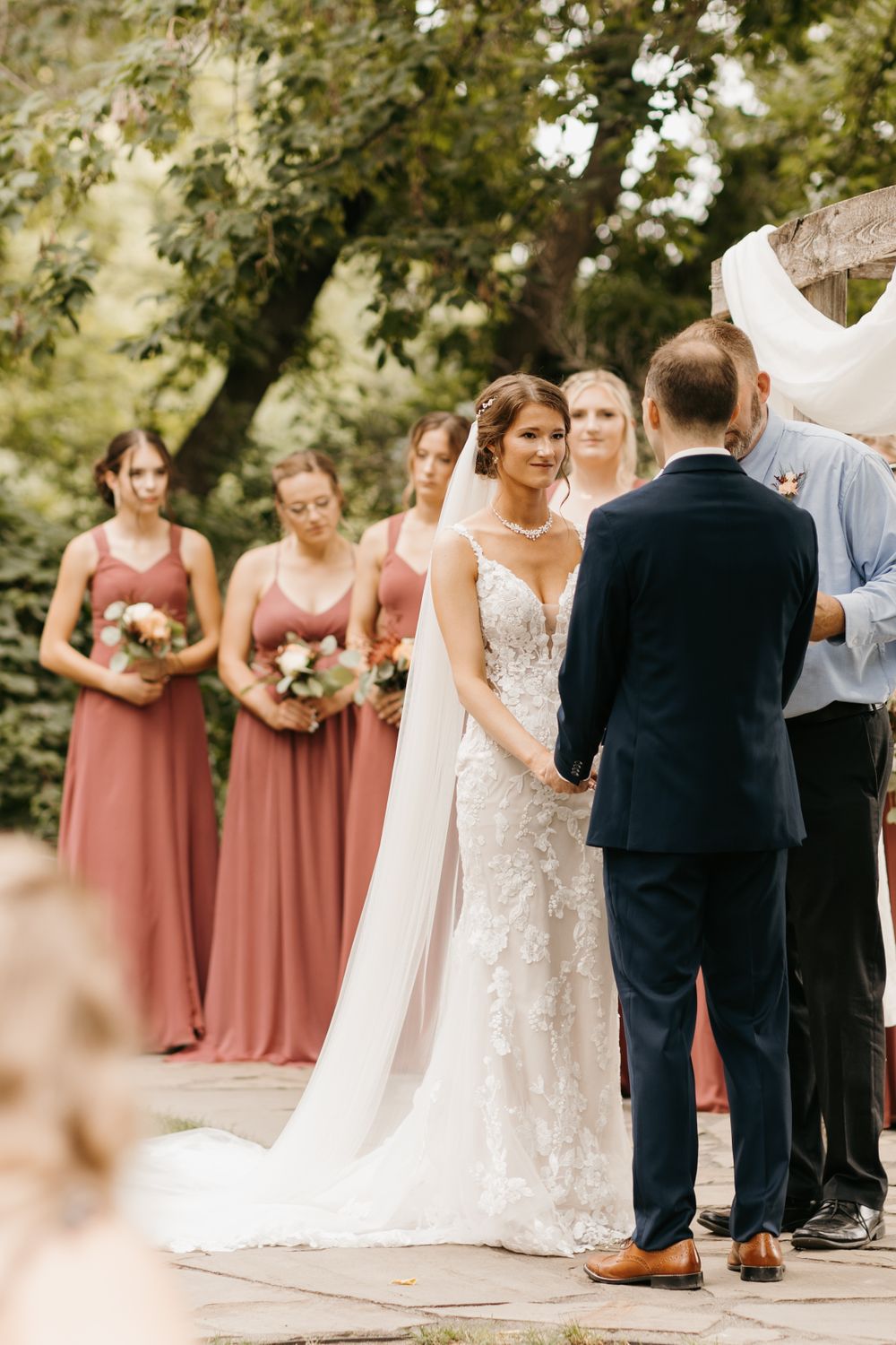 Bride in lace gown holds groom's hands at altar, bridesmaids in dusty rose dresses stand behind.