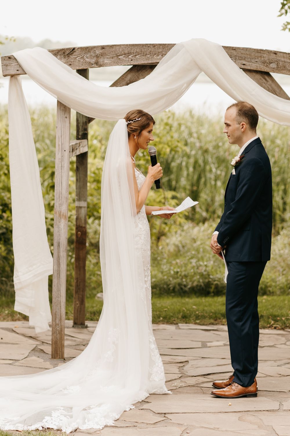 Bride reads personal vows into microphone at rustic wooden arbor draped with white fabric outdoors.