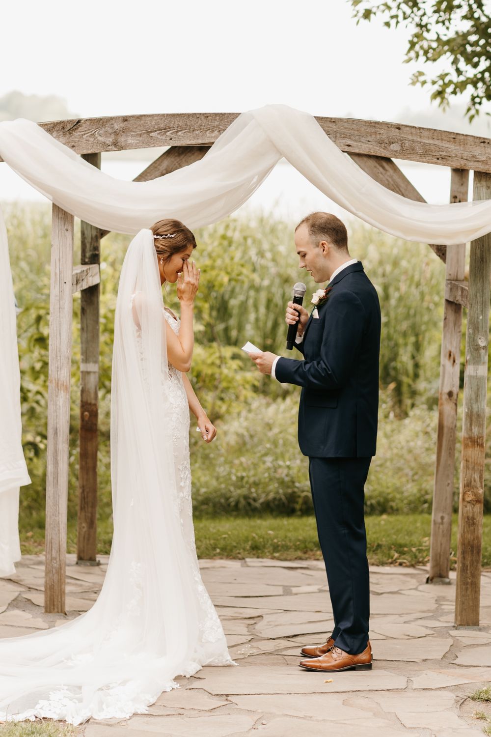 Emotional bride wipes tear as groom reads vows at rustic wooden arbor with white draped fabric.