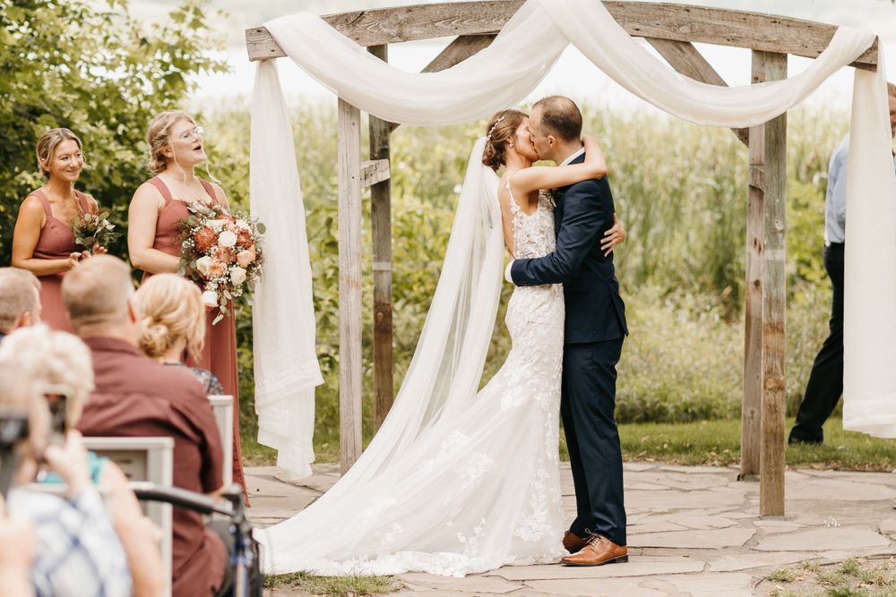 Bride and groom share first kiss under a rustic wooden arch with white draping at outdoor wedding.