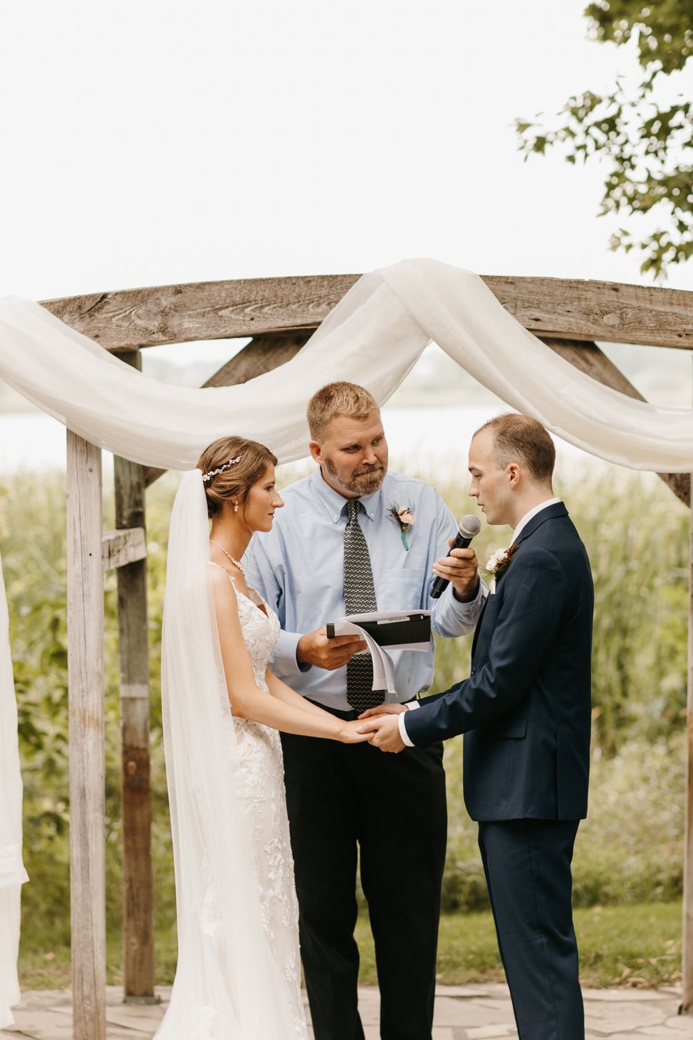 Officiant reads from book as bride and groom exchange vows under rustic wooden arbor with white draping.