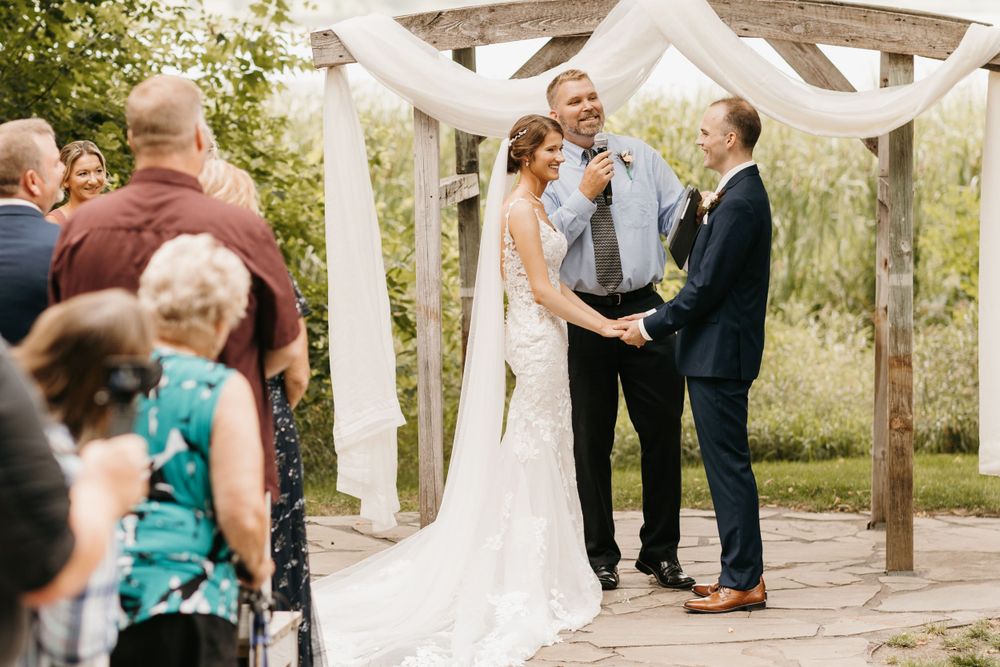 Bride and groom laughing with officiant during outdoor wedding ceremony under draped wooden arbor.