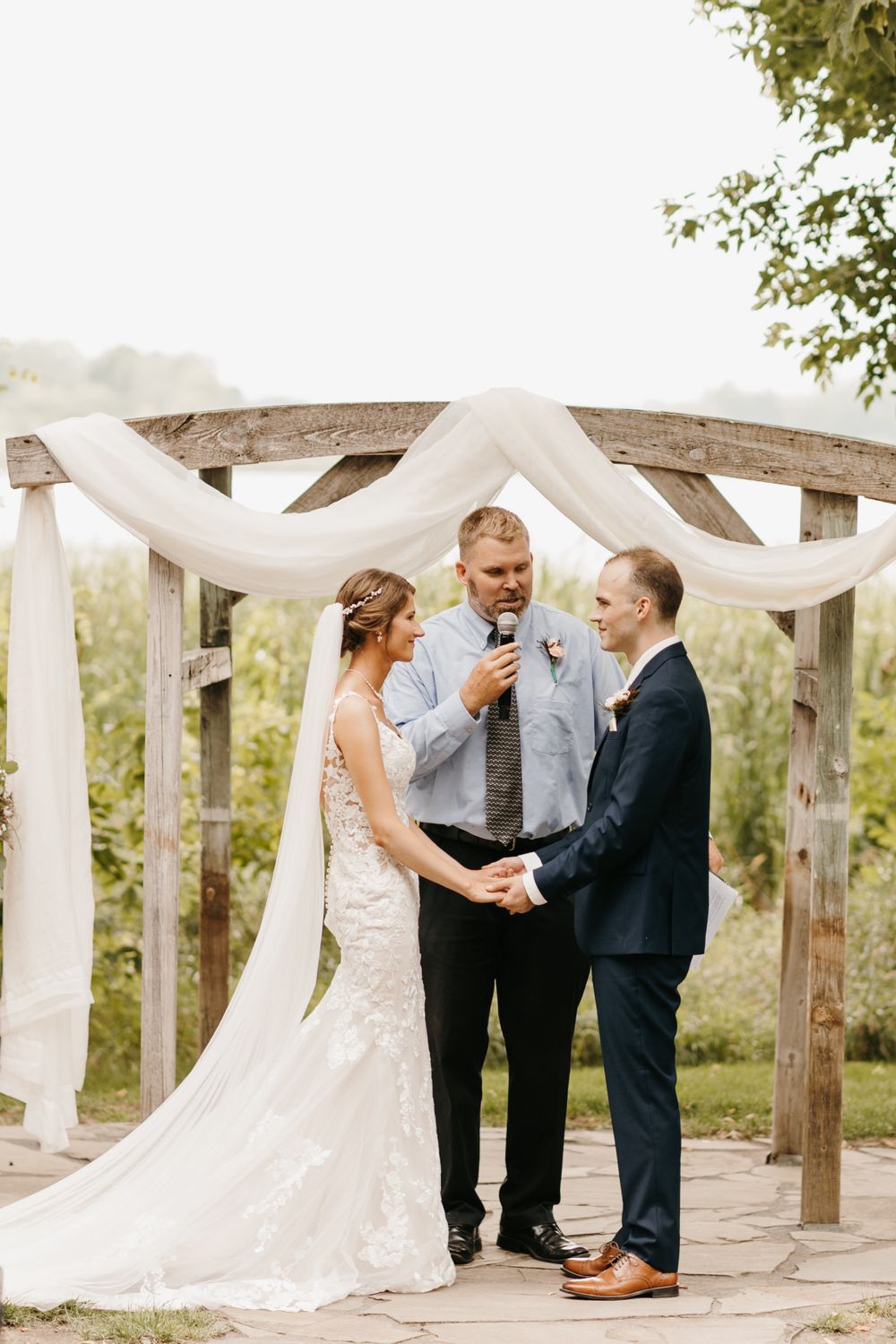 Bride and groom exchange vows at rustic wooden arch draped with white fabric during outdoor wedding ceremony.