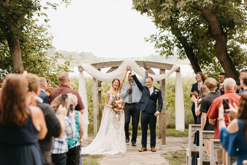 Newlyweds raise hands in celebration as they walk back down the aisle after outdoor wedding ceremony.