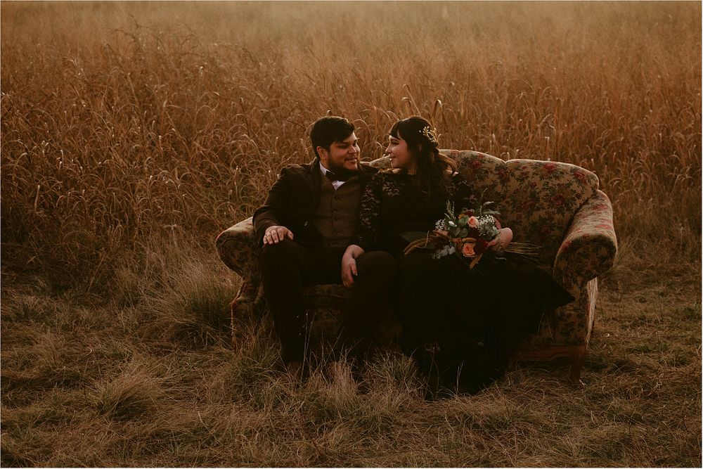 Couple in black attire sitting on vintage sofa in rustic field setting.