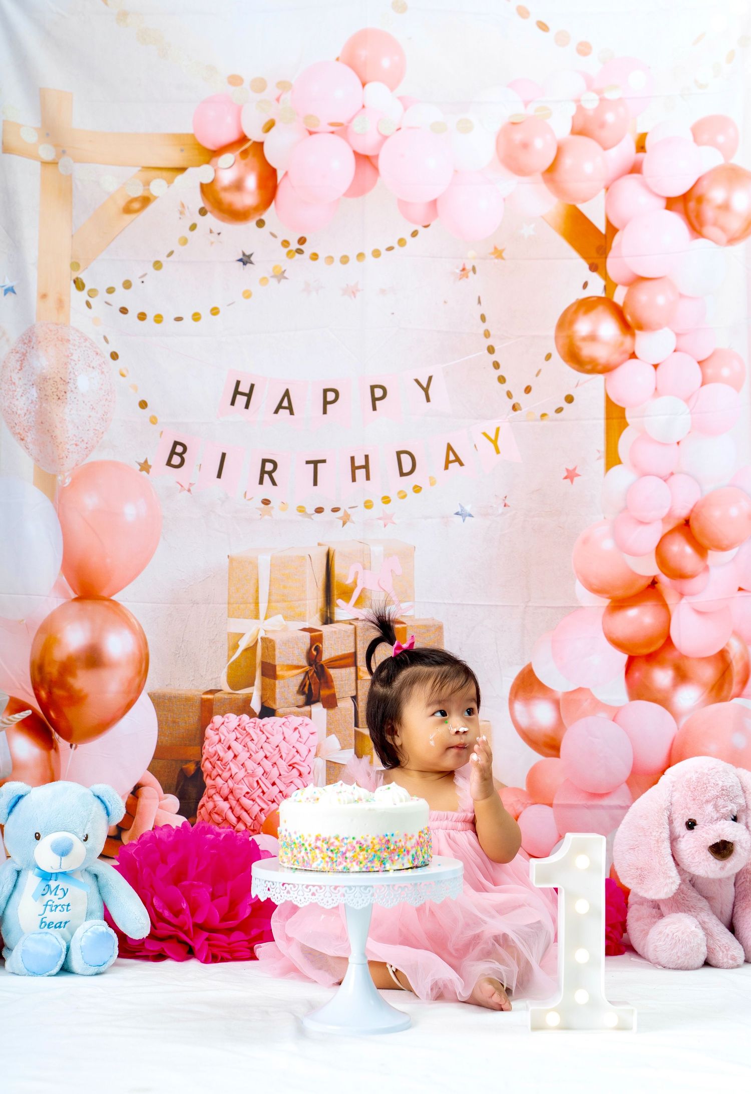 A festive first birthday setup with pink balloons, plush toys, gold gifts, and a birthday cake on a white backdrop.