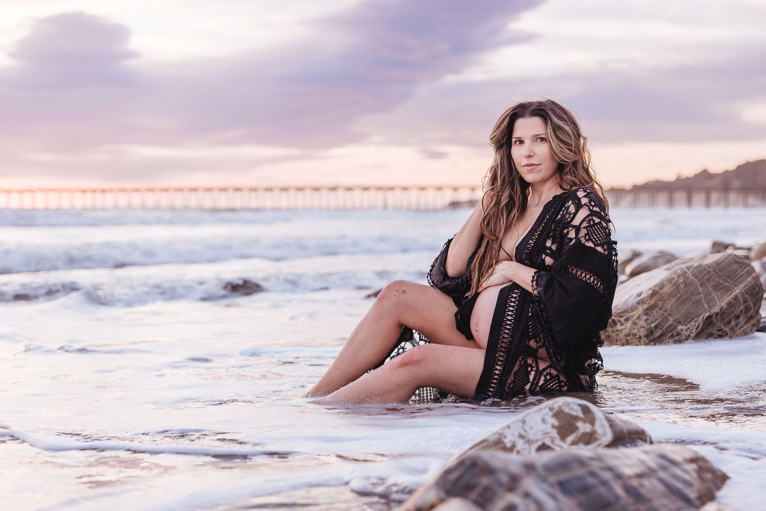 Woman in black lace dress relaxes on coastal rocks during golden hour.