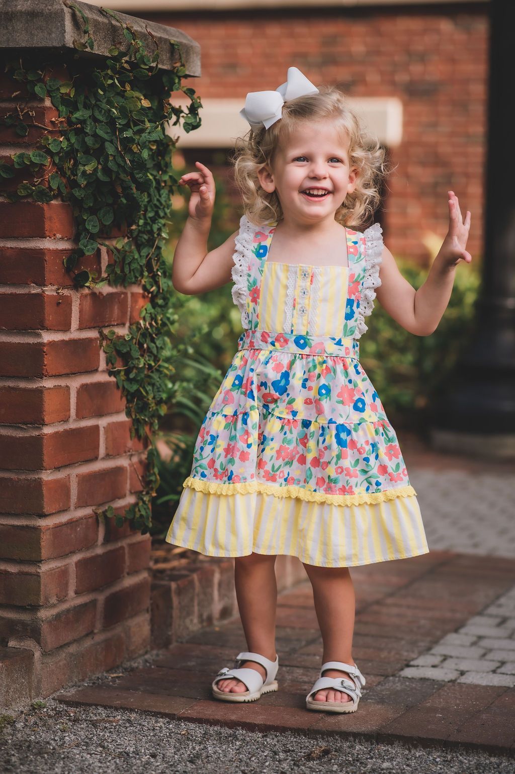 3-year-old in floral apron dress stands by ivy covered brick for milestone photo session
