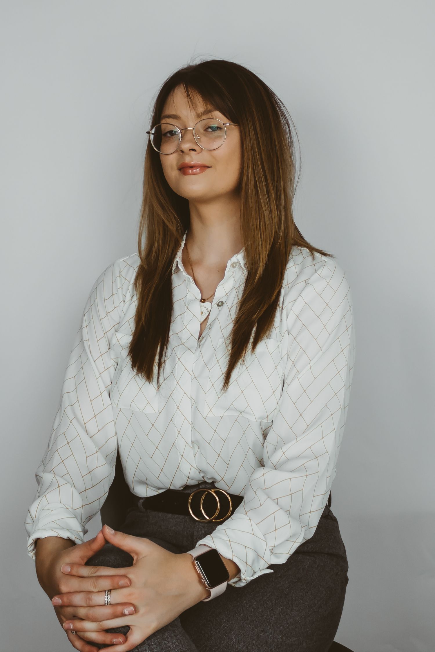 A portrait in white quilted blouse and brown skirt wearing round glasses against grey background.