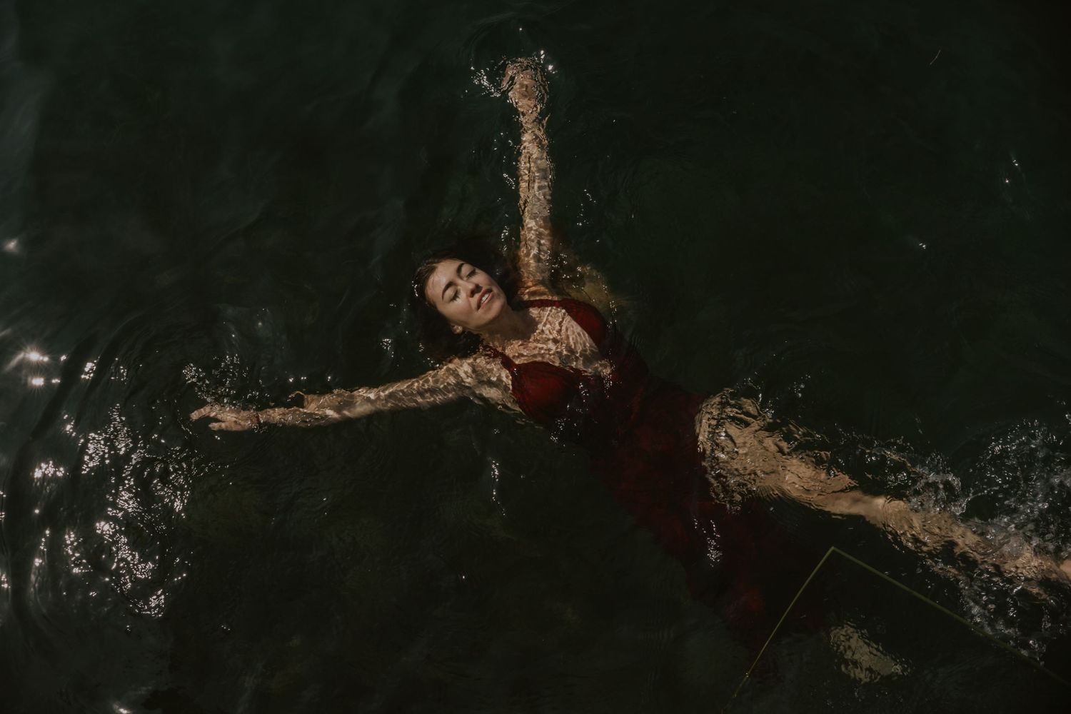 A figure in a red dress floats gracefully in dark water creating artistic ripples.