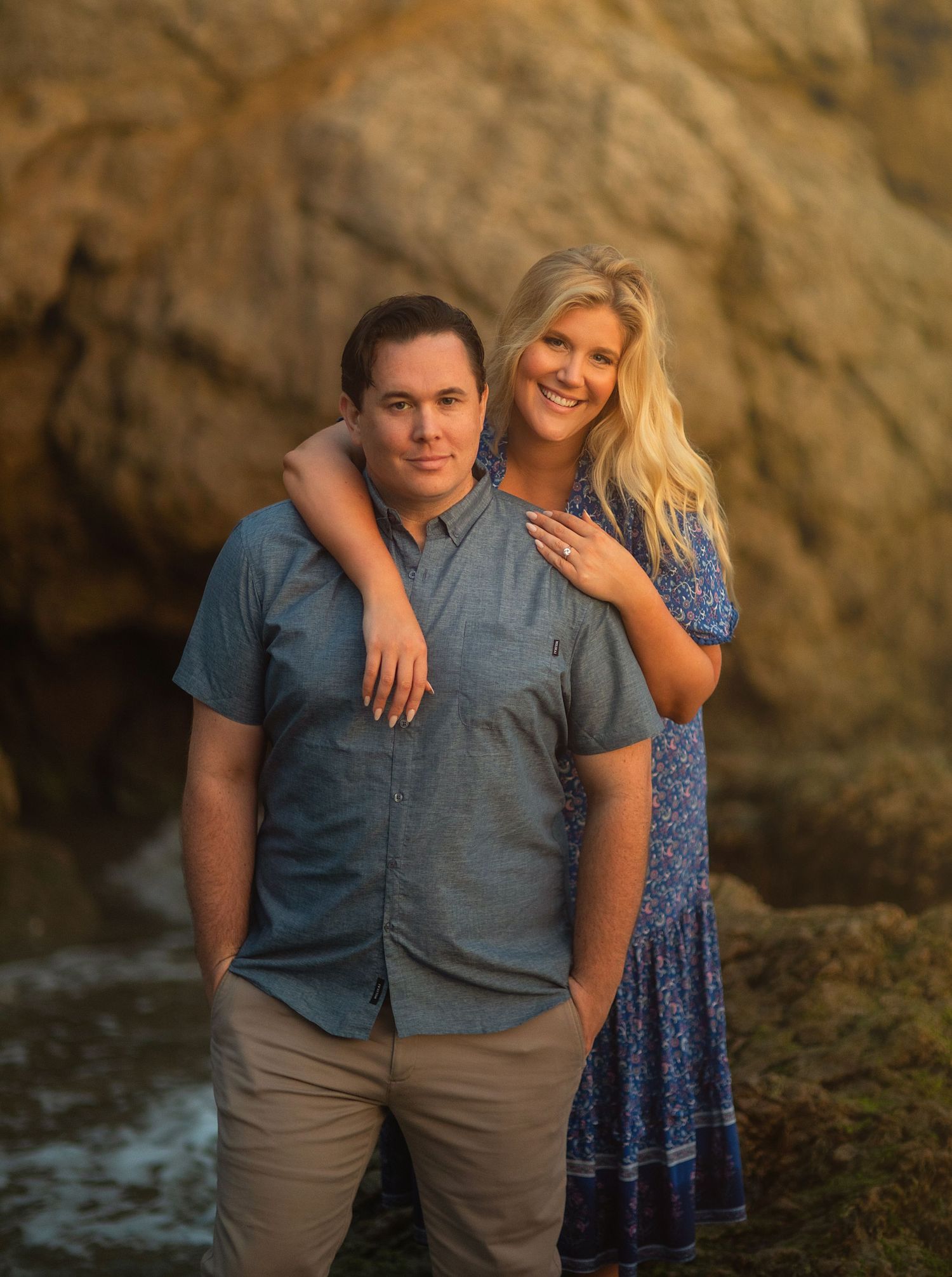 A couple poses together against a rocky backdrop near the ocean during golden hour.