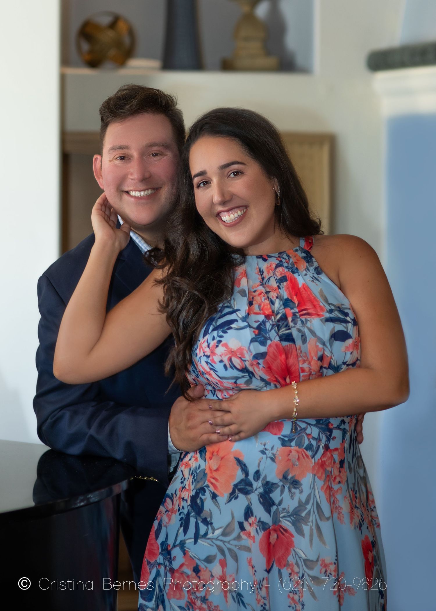 A couple smiles while posing together by a piano in their home.