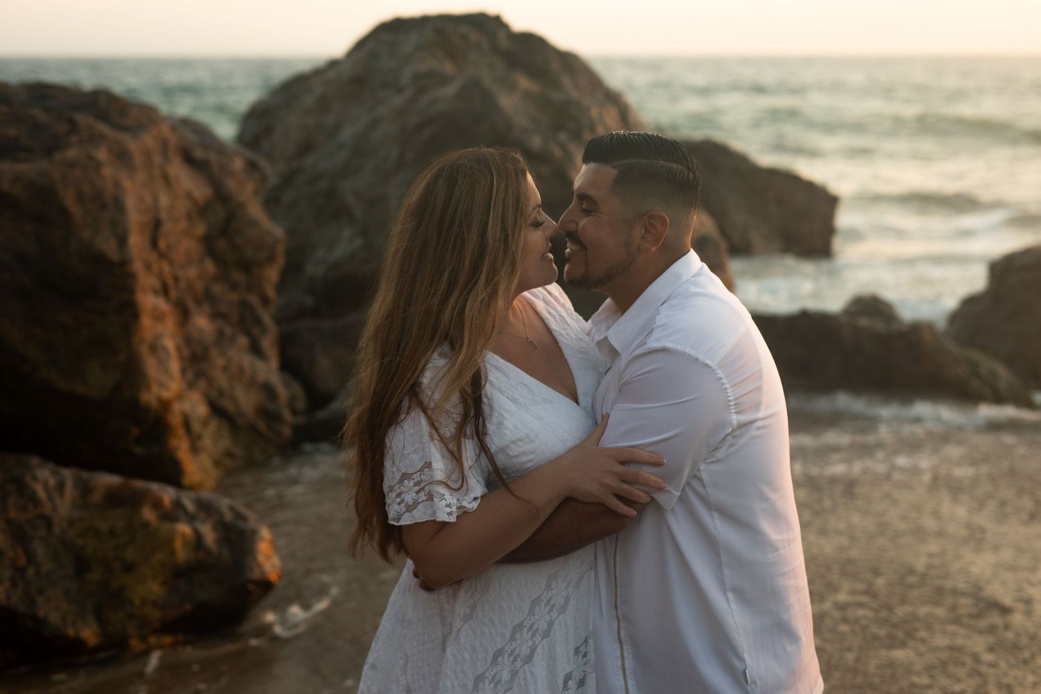 A romantic beach photo at sunset with rocky coastline in background.
