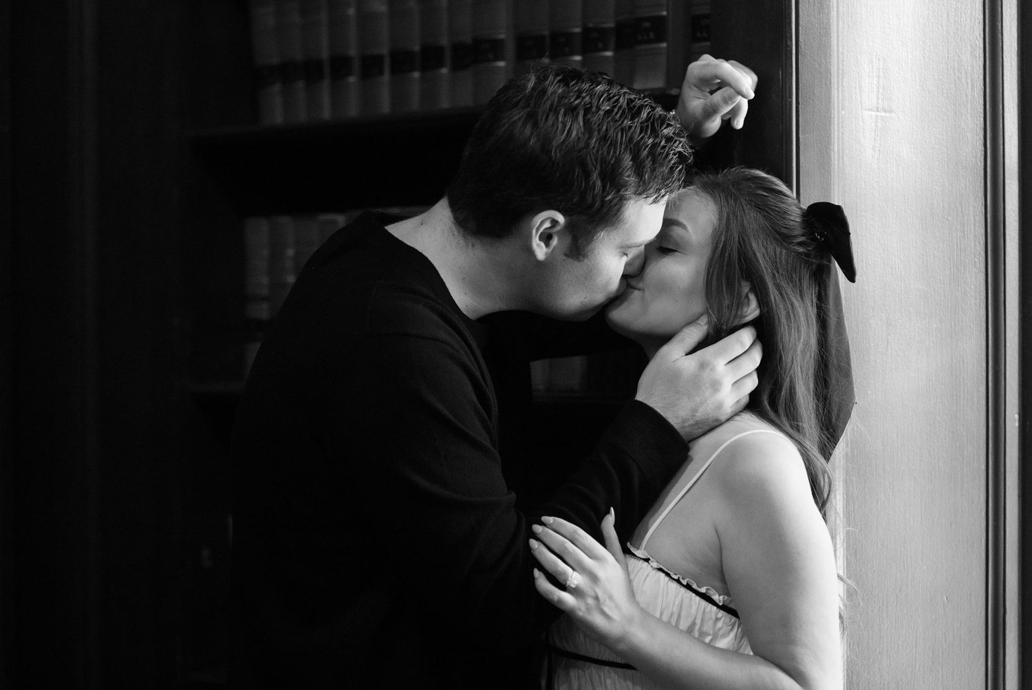 Black and white romantic portrait of a couple sharing an intimate kiss against a window.