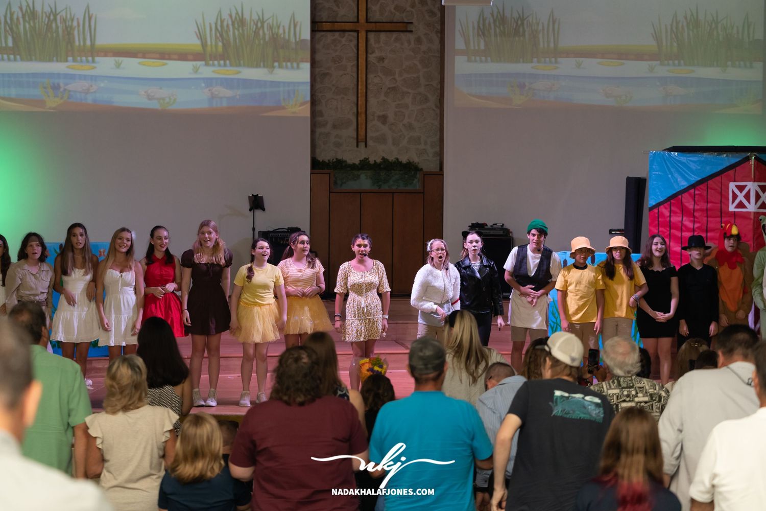 Large theater cast assembled on stage wearing various costumes under stage lighting.
