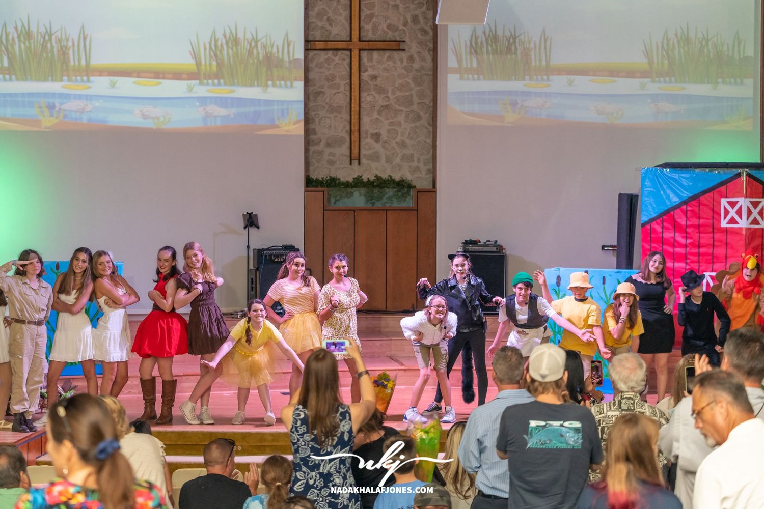 Full cast stands on stage during a theatrical performance with colorful lighting.