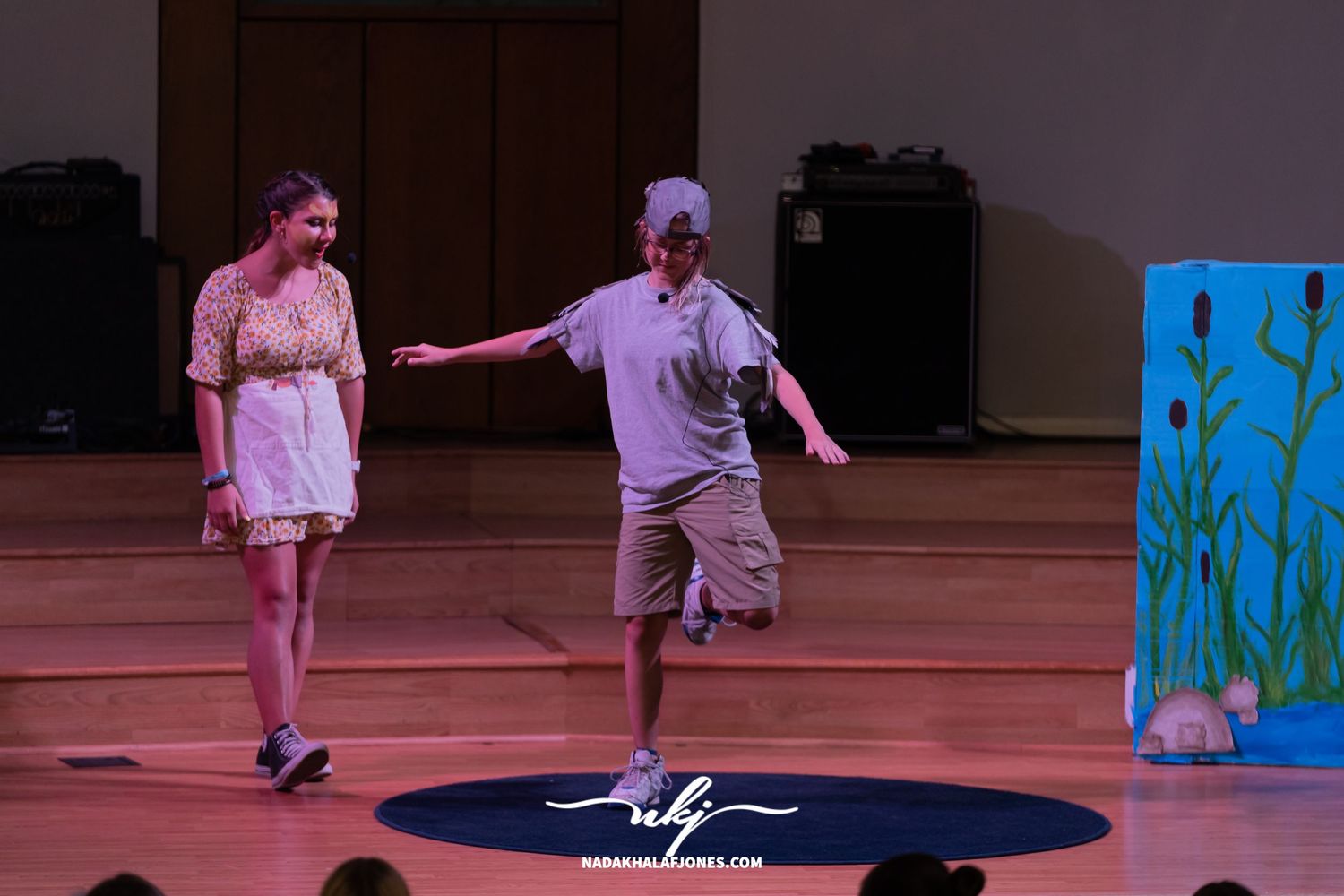 Theatrical performers move across a stage with dramatic lighting and scenic elements in the background.