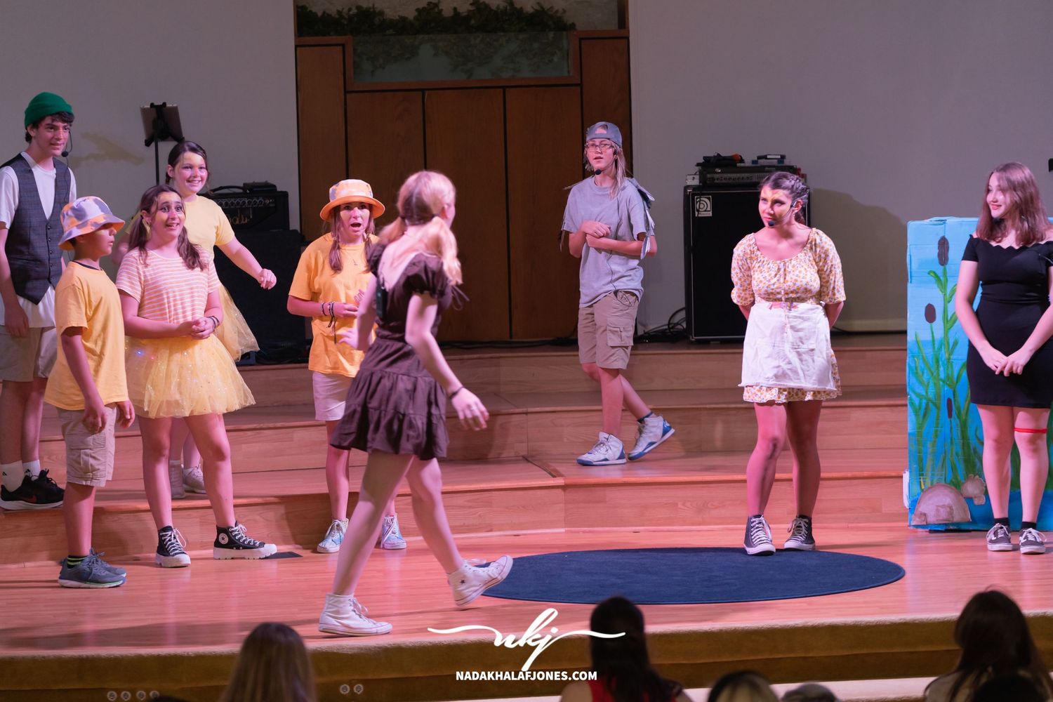 Theatrical performers in casual costumes act out a scene on a wooden stage.