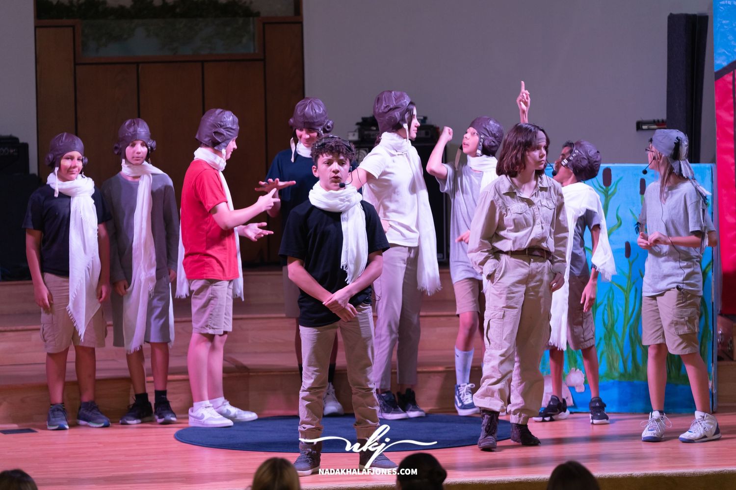 Theater production scene with actors in white outfits interacting on a stage with blue lighting effects.