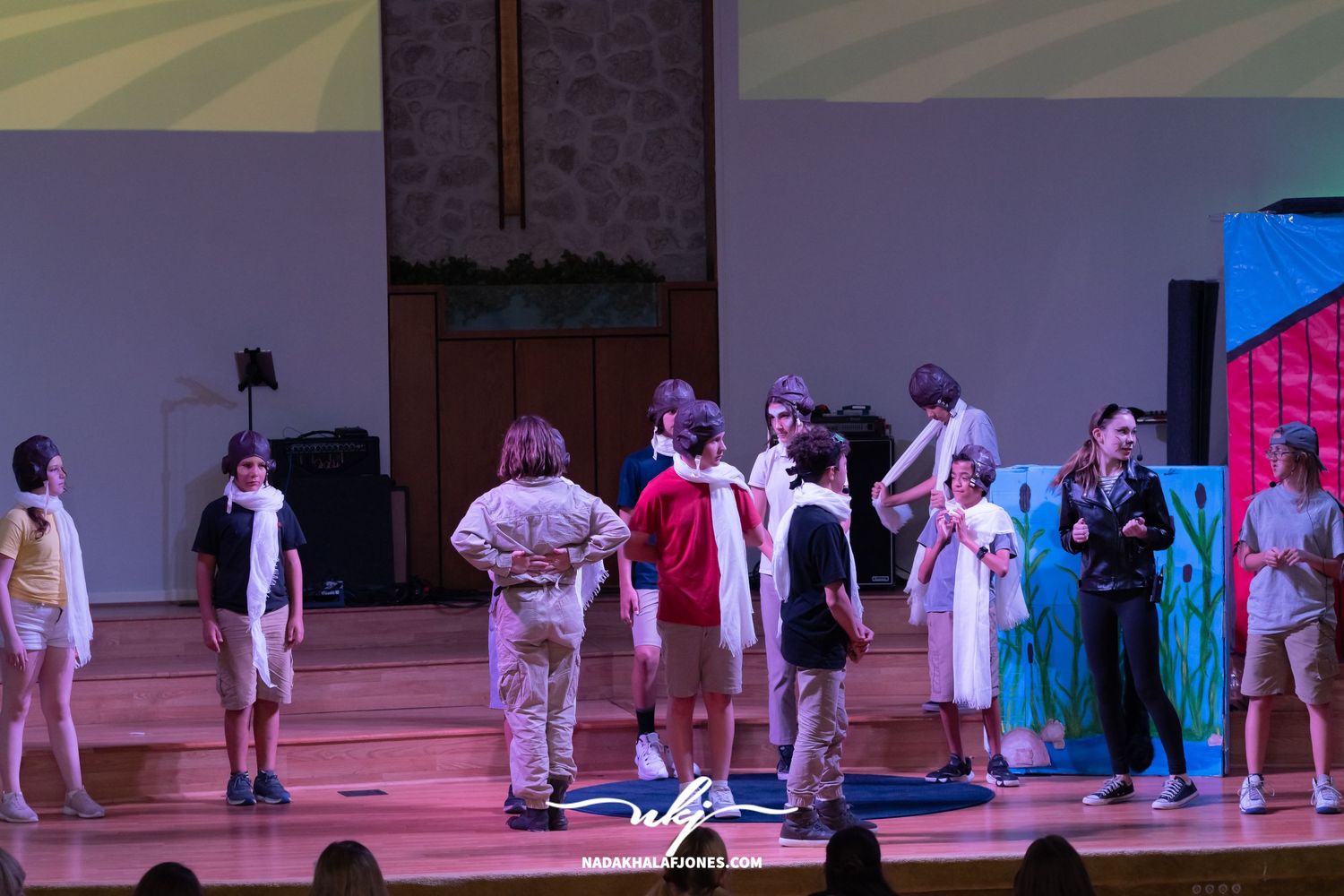 Theatrical scene with performers arranged across stage featuring scenic backdrop and colored lighting design.