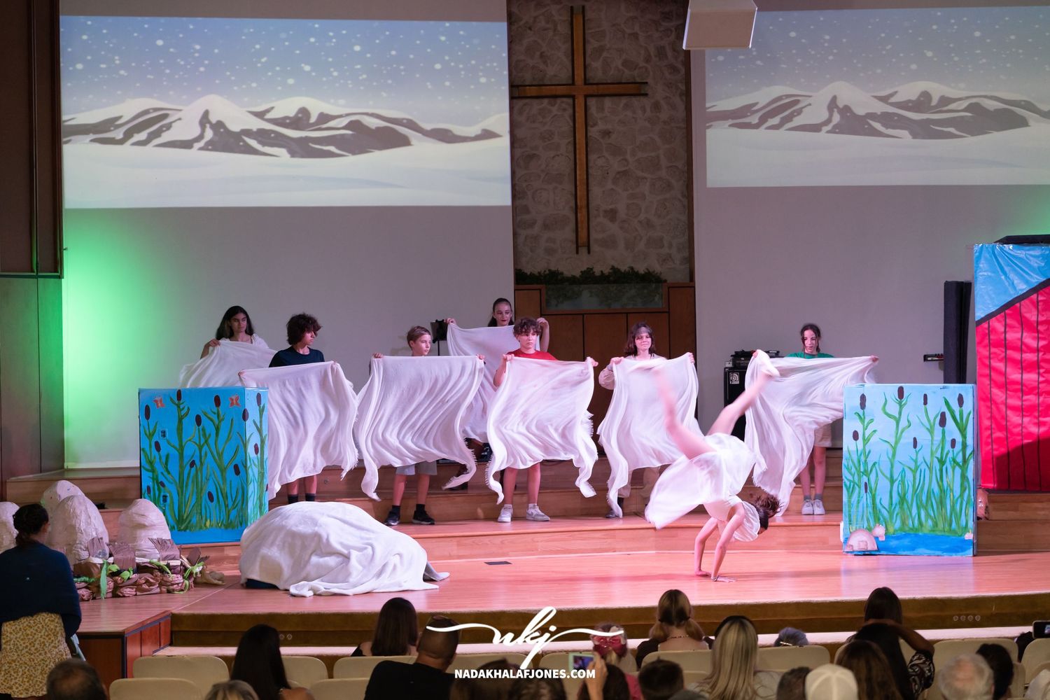 Theater stage with mountain backdrop and performers arranged in dramatic formation under stage lights.