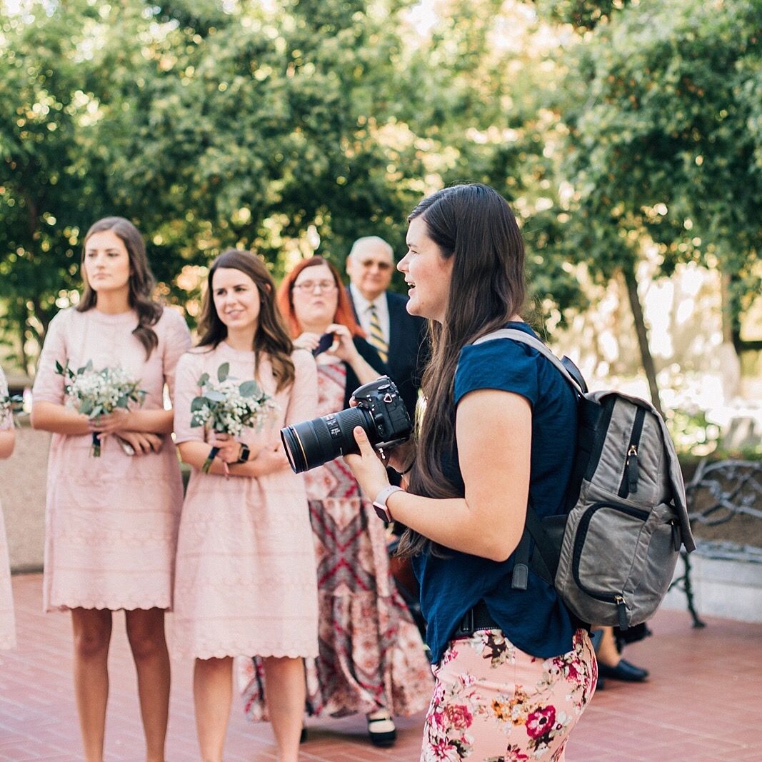 Photographer in navy top captures bridesmaids in pink dresses holding bouquets during outdoor wedding.