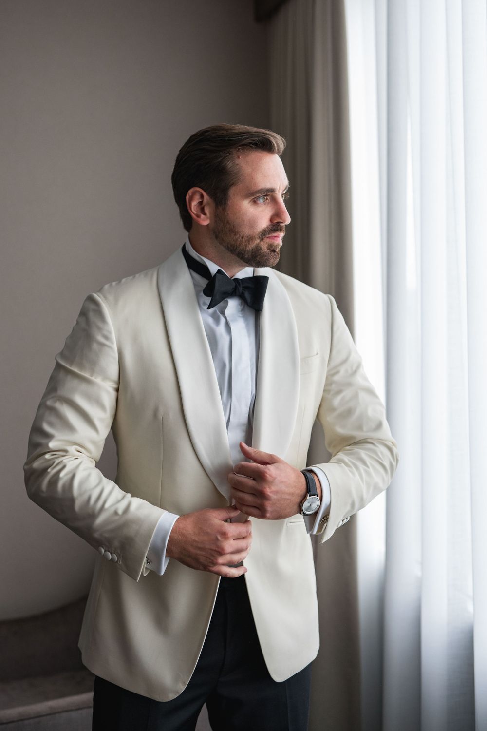 A groom in a white tuxedo jacket and bow tie gazes out a window.