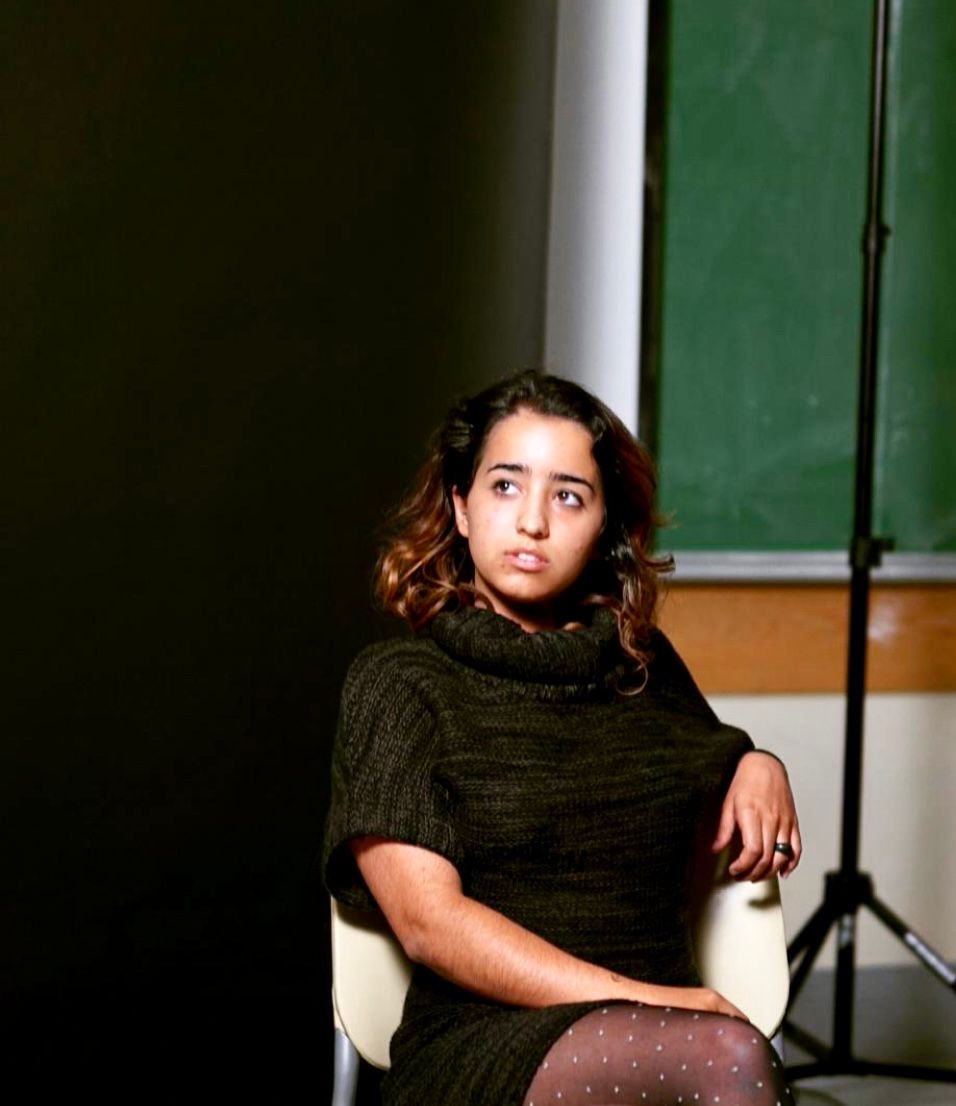 Woman with dark hair and eyes, olive complexion looks to her right, seated against a dark background
