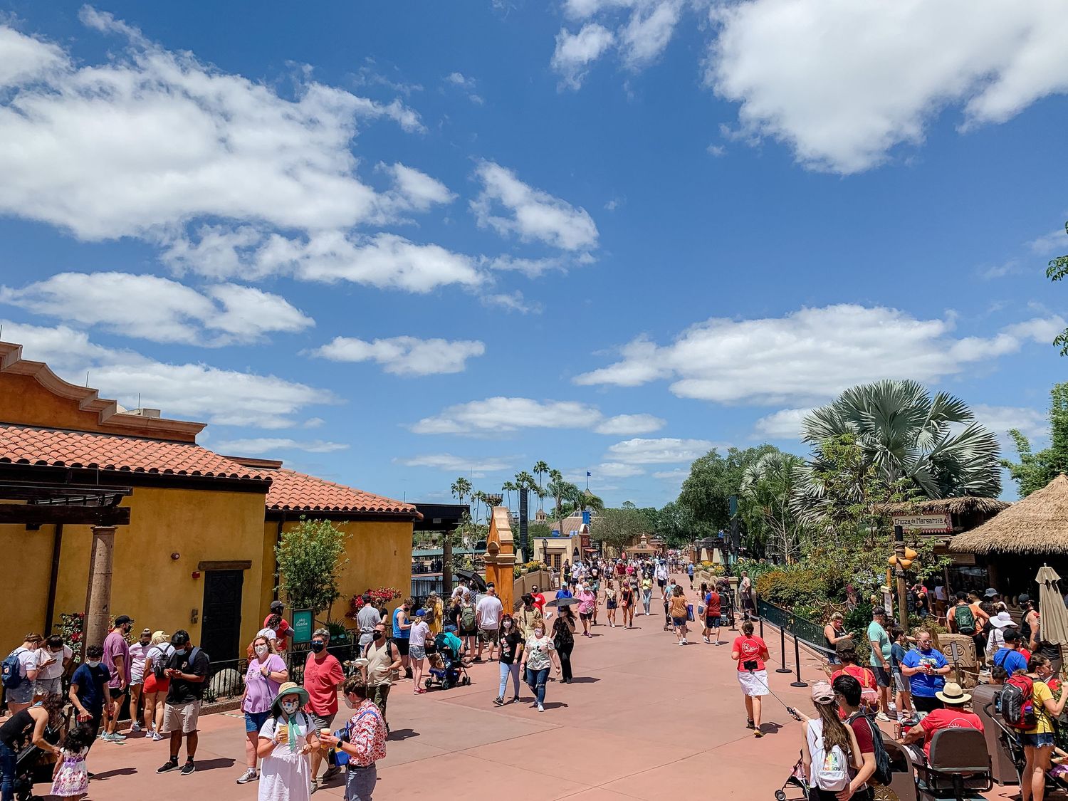 Crowded walkway in a theme park with Spanish-style buildings and palm trees on a sunny day.