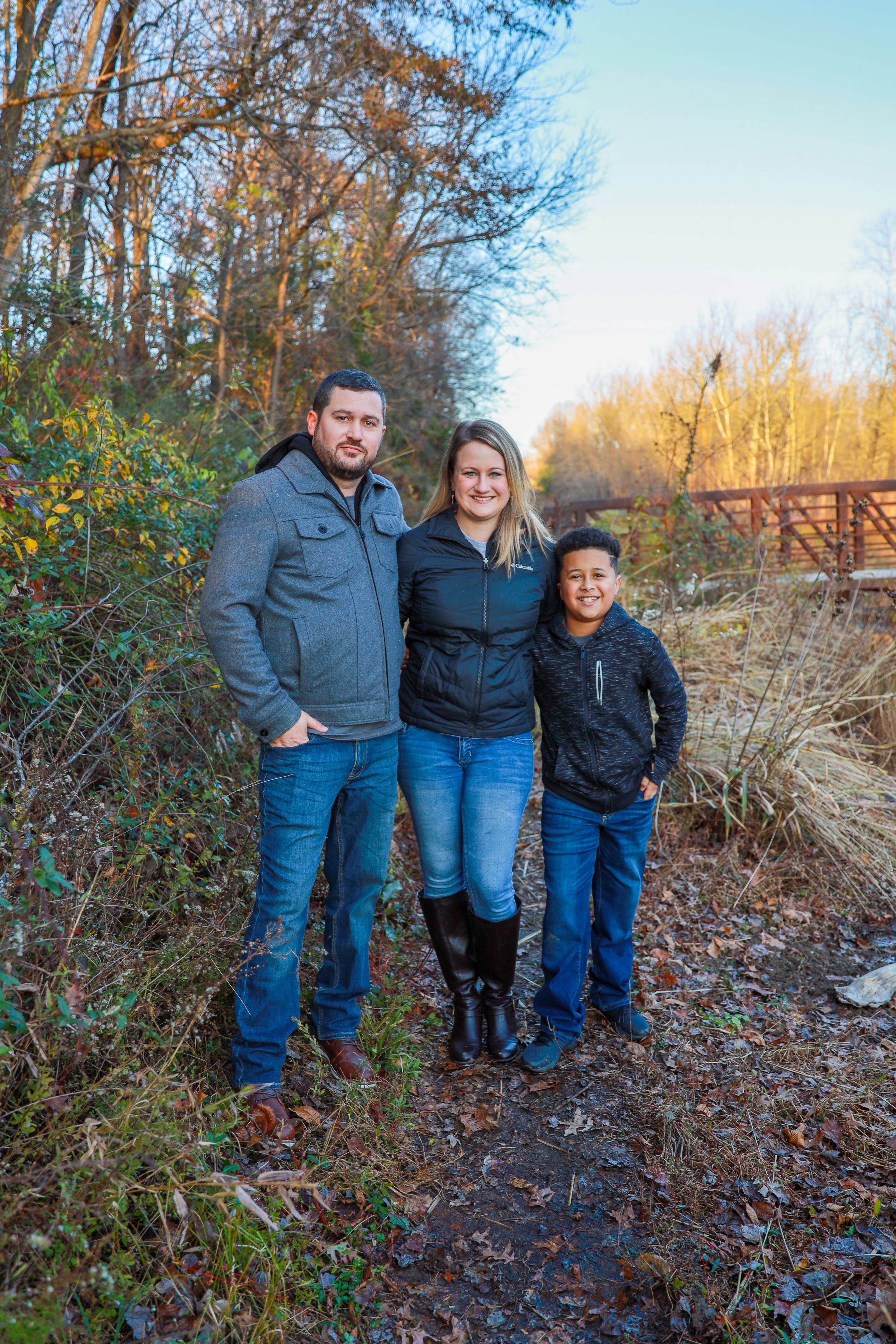 Three people pose together on a fall nature trail surrounded by dried brush and leaves.