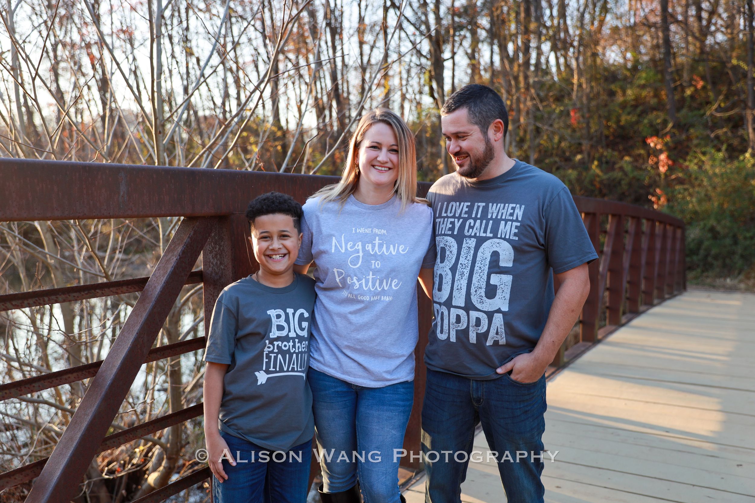 A family photo session captured in golden autumn sunlight on a wooden bridge with fall foliage in the background.
