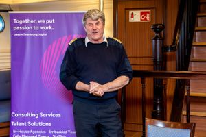 Speaker stands at a corporate consulting services presentation podium with purple branding banner in background.