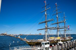 Tall sailing ship with multiple masts docked at harbor against bright blue sky.