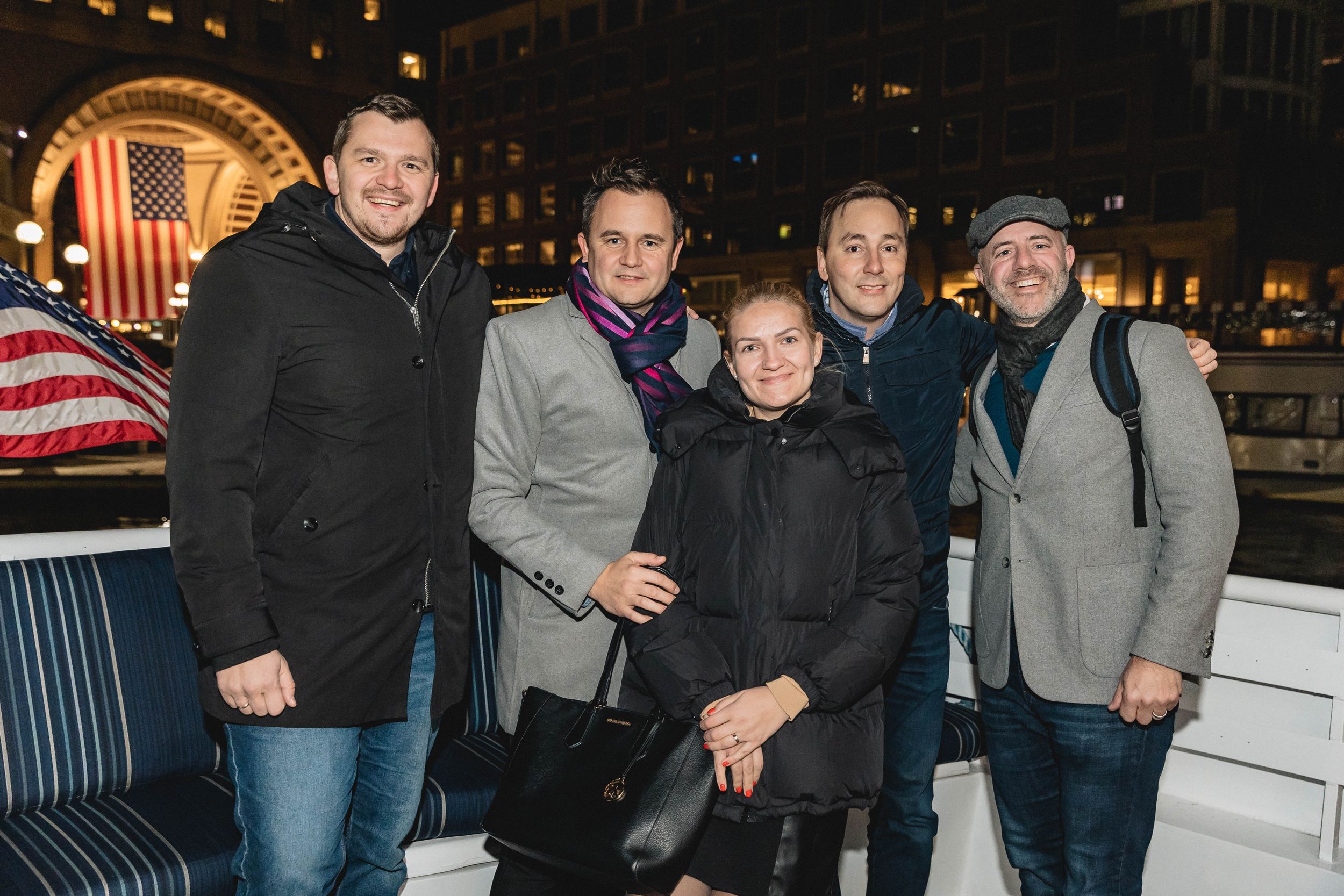 Small group of people bundled in winter coats posing together at night with American flag visible.