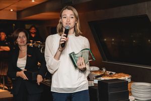 Speaker holds a microphone while presenting at an indoor event with tables and guests in the background.