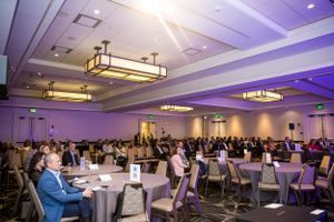 Large conference room filled with attendees at round tables under modern lighting fixtures.