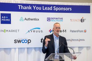 Speaker presents at podium in front of corporate sponsor wall displaying various tech company logos.