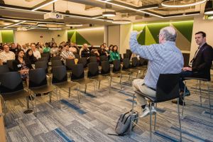 Speaker presenting to large audience in modern office space with geometric wall design.