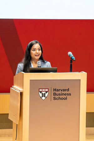 Speaker presents at Harvard Business School podium with red backdrop and school shield.