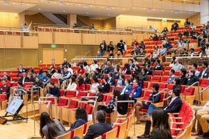 Audience members seated in tiered red chairs at a professional conference venue.