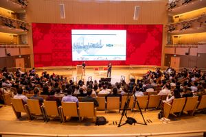 Wide view of a packed auditorium with red stage backdrop and projection screen during a presentation.