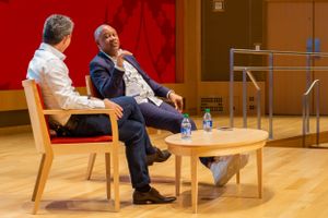 Two speakers engage in a discussion on stage with wooden chairs against a red backdrop at a professional event.