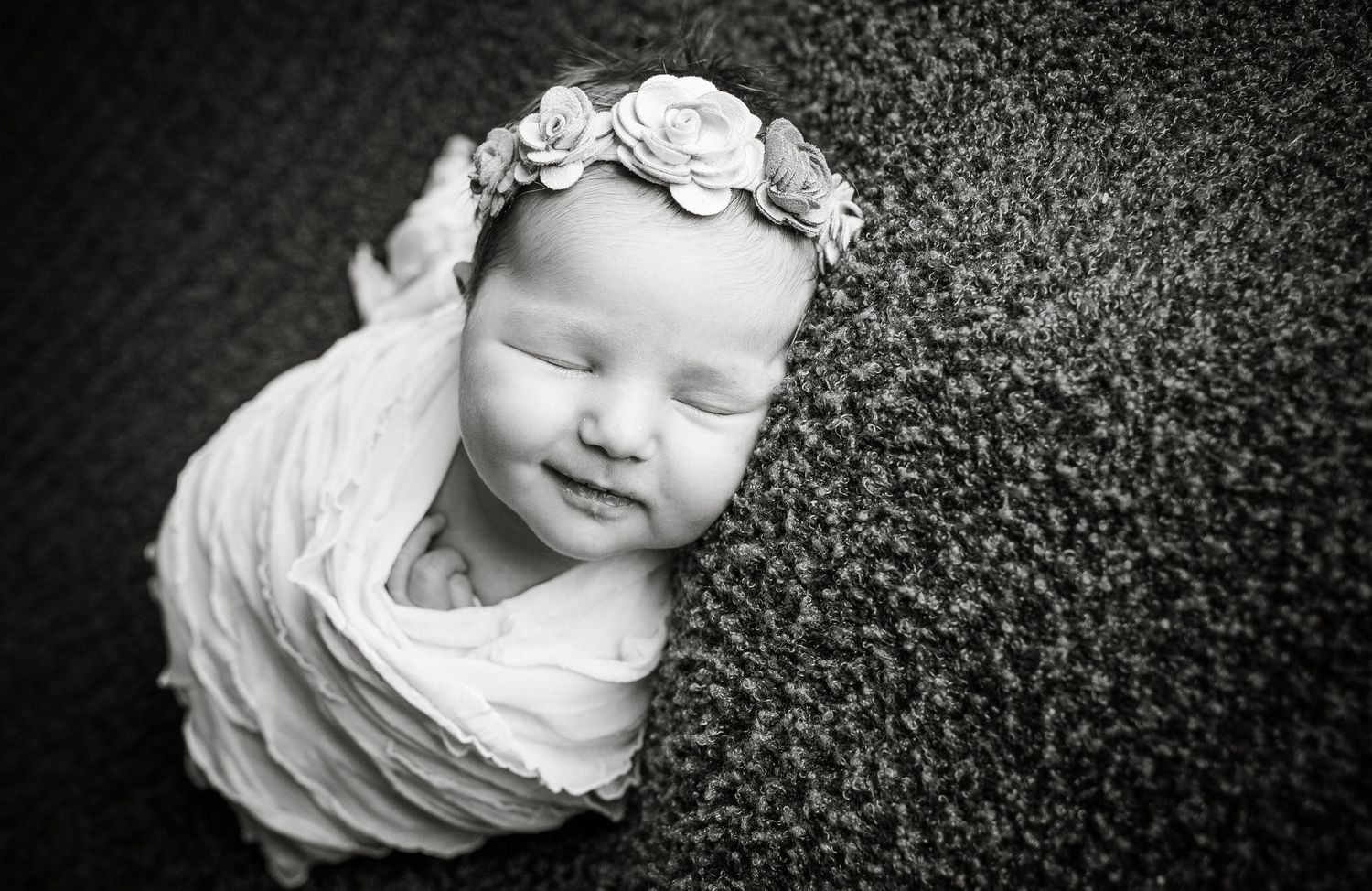 Black and white portrait of a newborn wrapped in white fabric wearing a delicate floral headband on textured background.