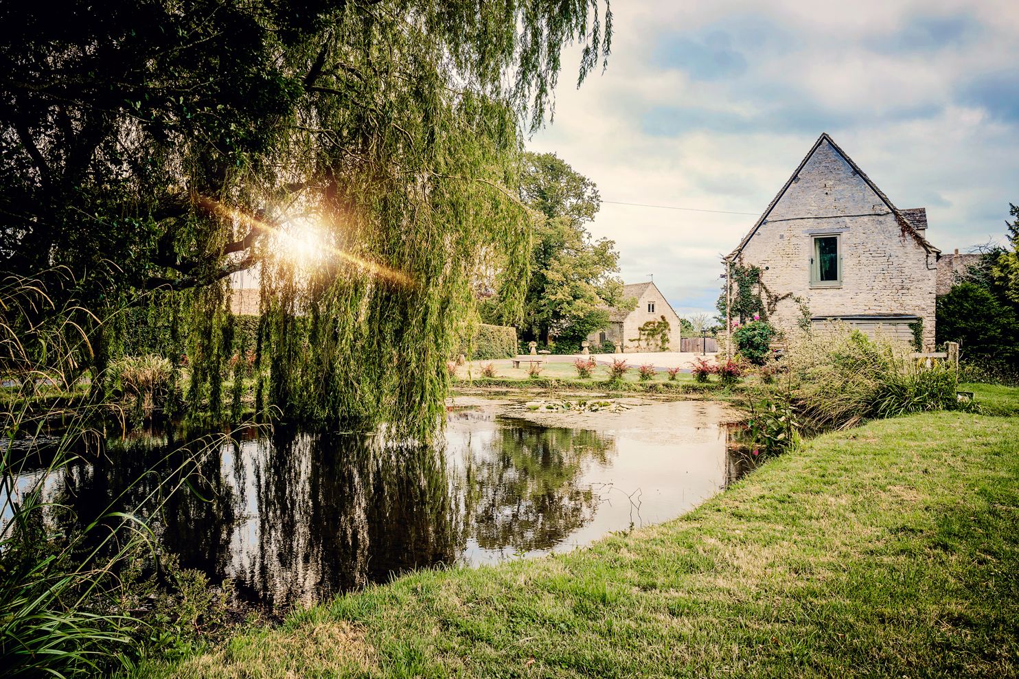 Rustic white house beside a peaceful pond with weeping willow tree and sunburst through branches.