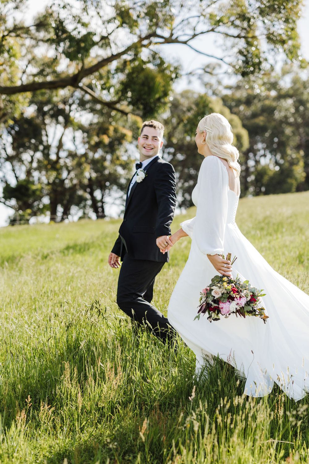 Newlyweds walk through tall grass with bride holding a burgundy bouquet.