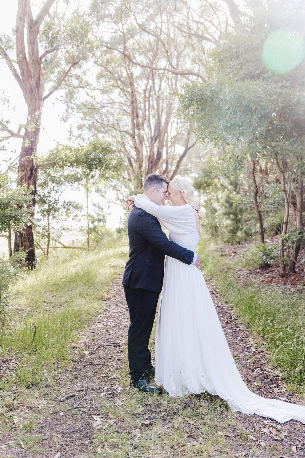 Romantic wedding moment captured on a tree-lined path in natural sunlight.