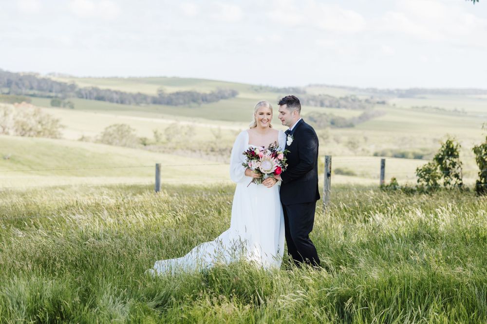 Couple in wedding attire standing in a scenic grassy meadow with rolling hills in the background.