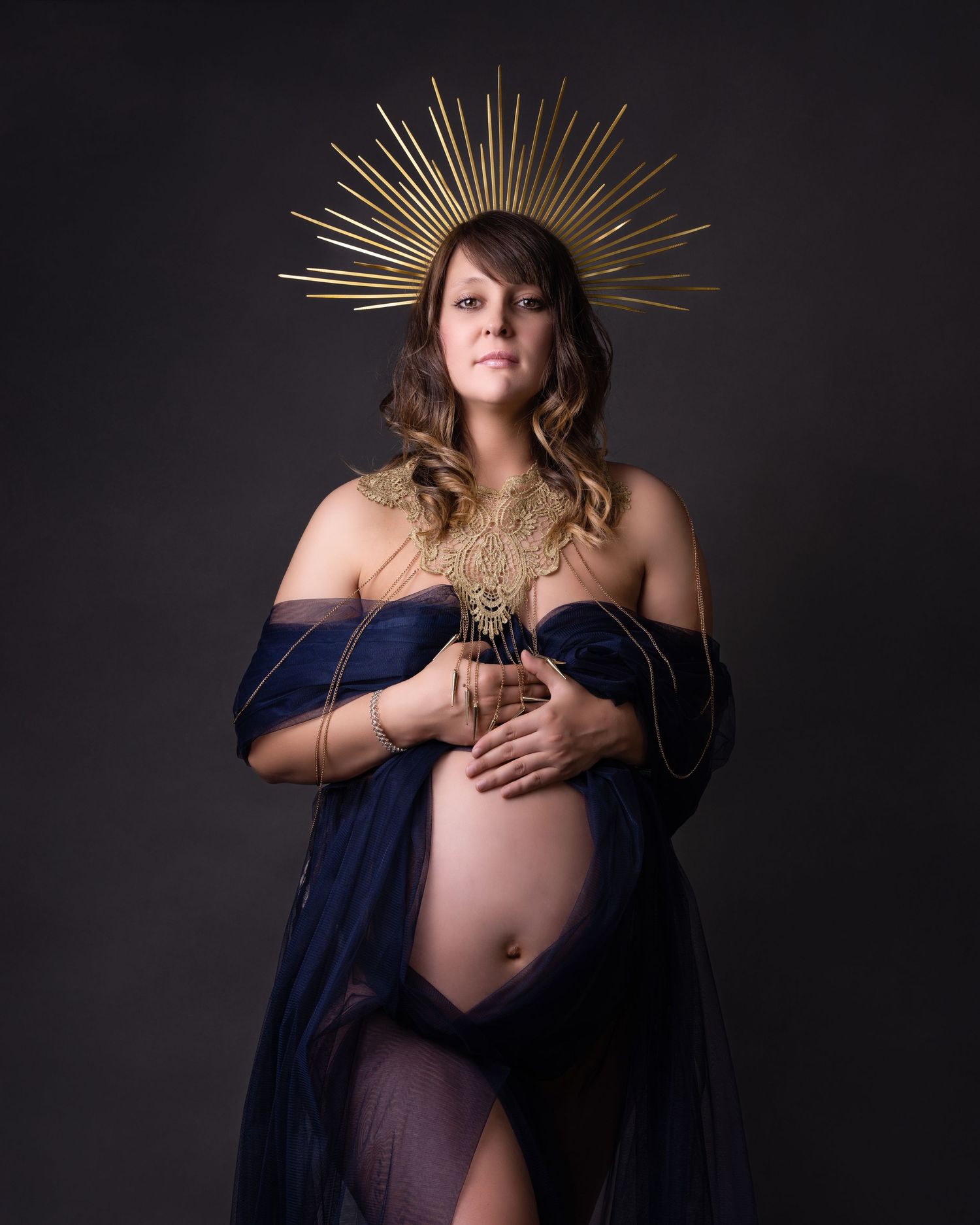 Artistic maternity portrait featuring navy fabric and gold sunburst crown against dark background.
