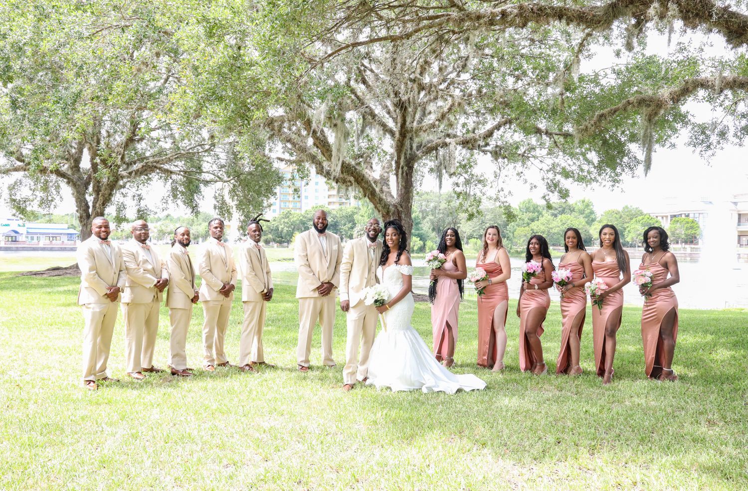 Wedding party in beige and pink attire posed outdoors under trees.