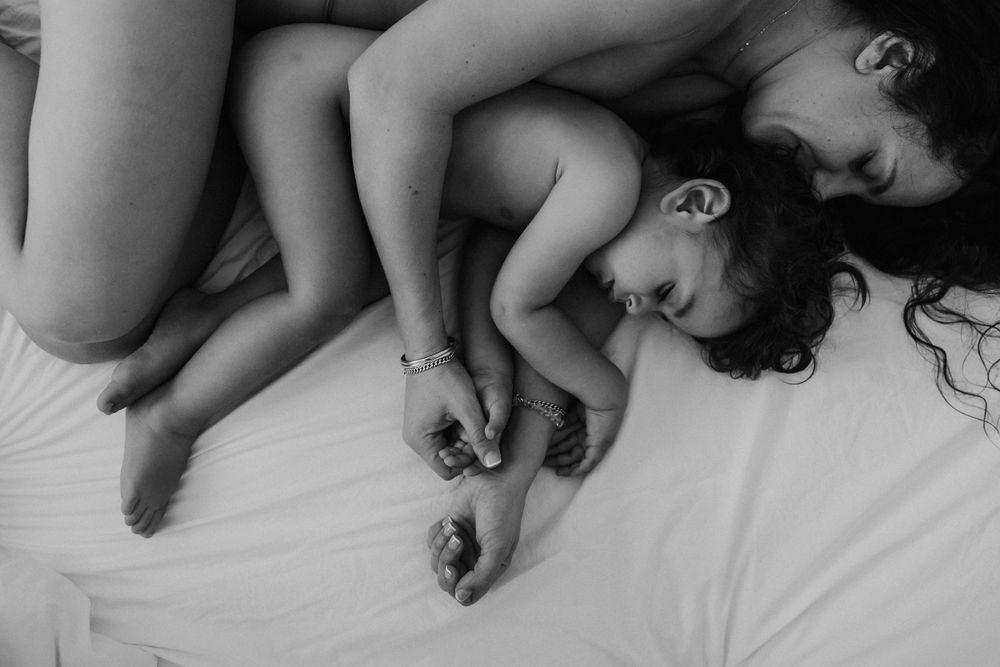 Black and white intimate photo of mother and curly-haired toddler sleeping peacefully together on white sheets.